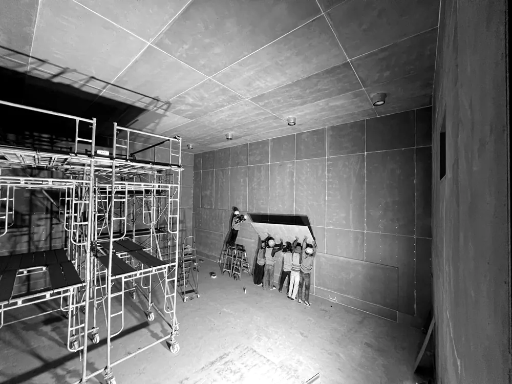 Walls and ceiling lined with layers of board, with visibly offset joints, as installers work at the rear of the room